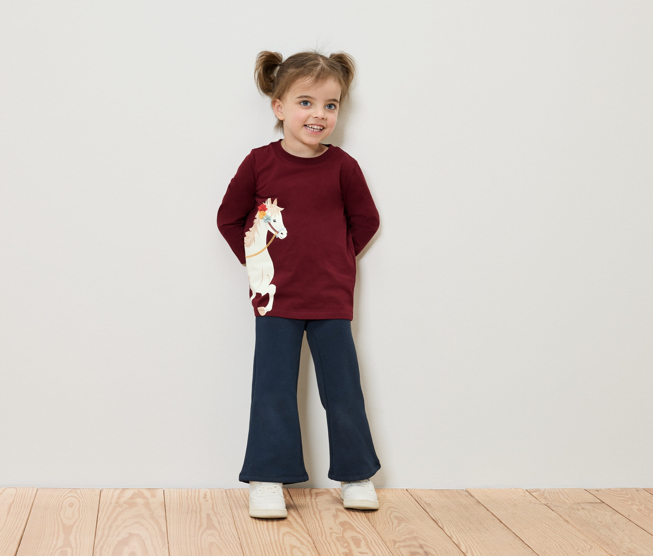 Uśmiechnięta dziewczynka z kucykami stoi przy ścianie, ubrana w bordową koszulkę z koniem i niebieskie spodnie.
