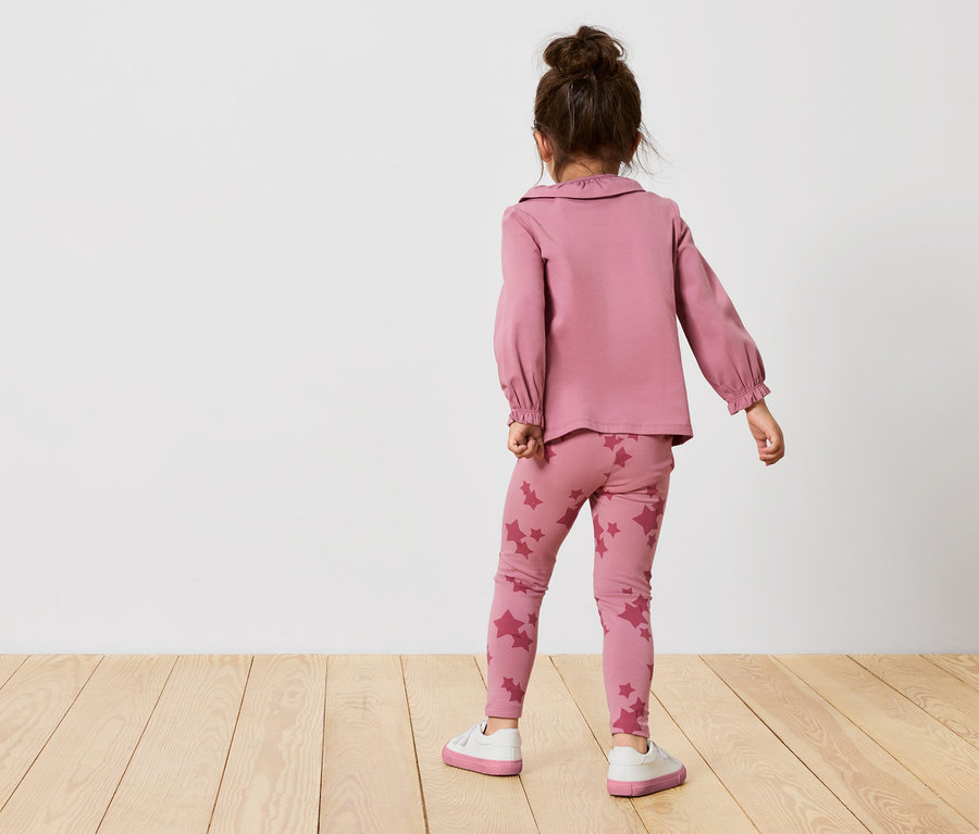 Dziewczynka w różowym komplecie w gwiazdki stoi tyłem. Ma na sobie różową bluzkę z długim rękawem i legginsy w gwiazdki.