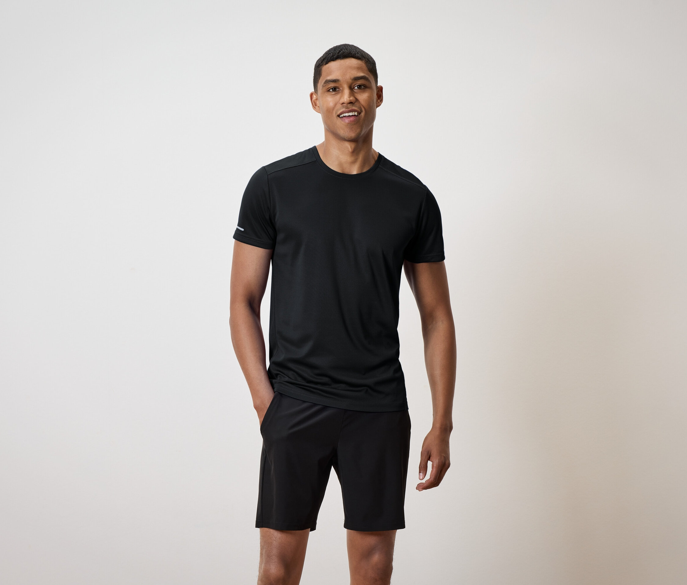 Uśmiechnięty mężczyzna stoi z rękami w kieszeniach czarnych szortów i ma na sobie czarną koszulkę.