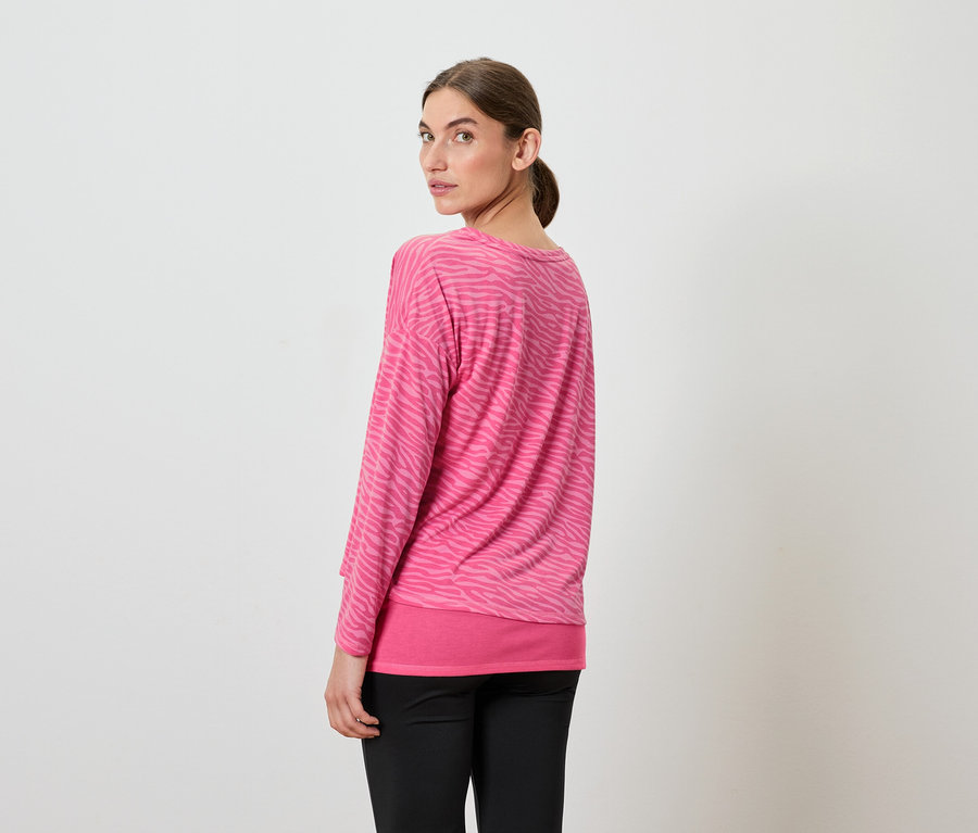 Kobieta patrzy przez ramię, ma na sobie różową bluzkę z długim rękawem i czarne spodnie.