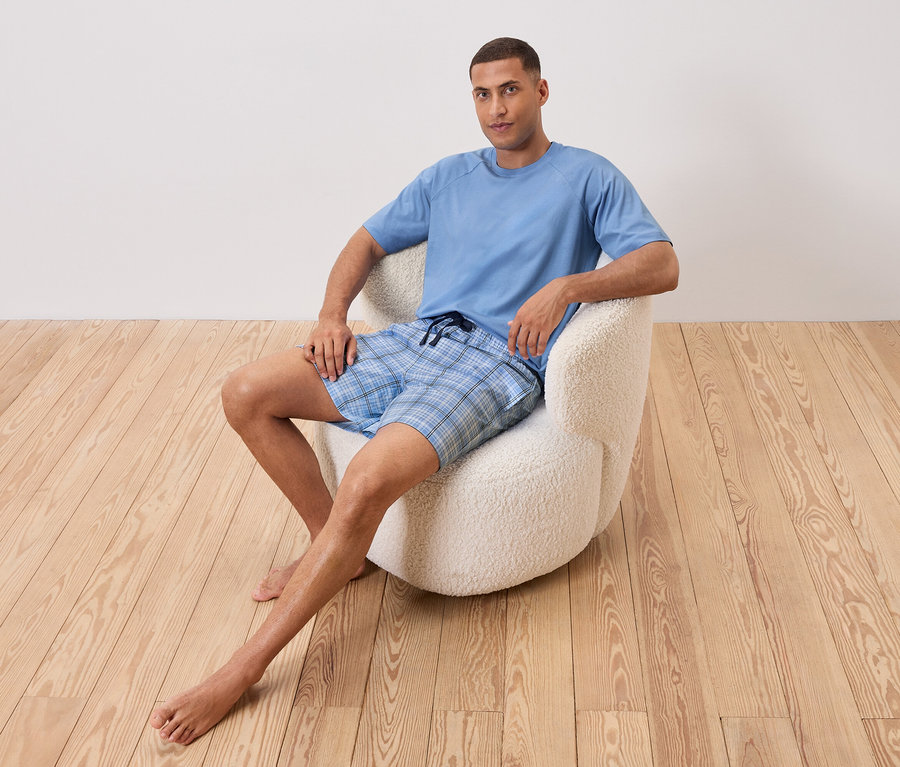 Mężczyzna siedzi boso w białym fotelu. Ma na sobie jasnoniebieski T-shirt i szorty w niebieską kratę.