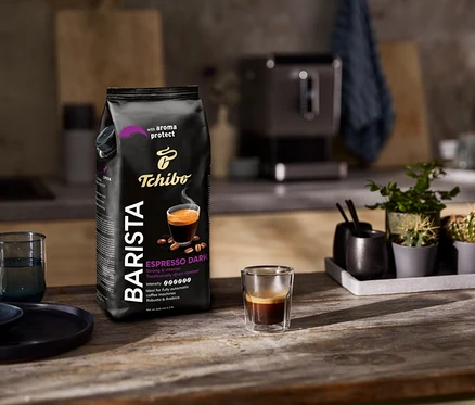 Paczka kawy ziarnistej Tchibo Barista Espresso Dark stoi obok szklanki espresso na drewnianym stole.