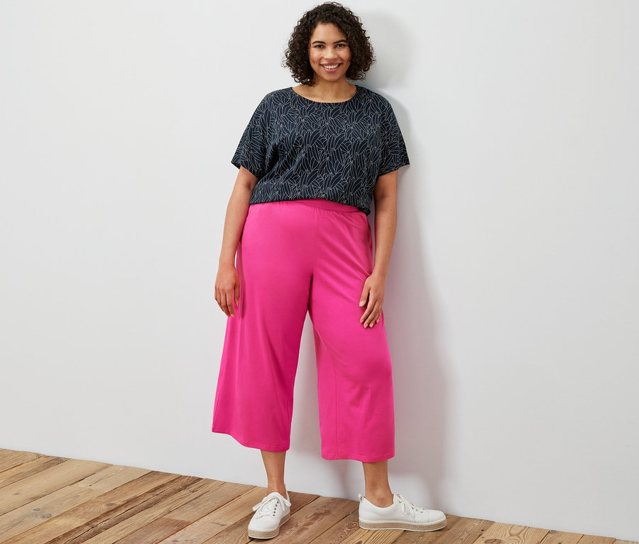 Kobieta stoi i ma na sobie granatową bluzkę z krótkim rękawem z obniżoną linią ramion i różowe spodnie kuloty z dżerseju.