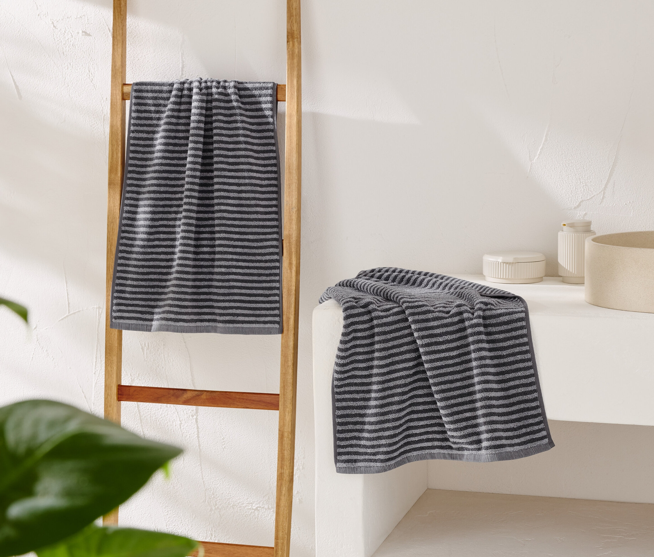 Pasiasty szary ręcznik wisi na drewnianej drabince, a drugi leży na białej toaletce obok beżowej umywalki.