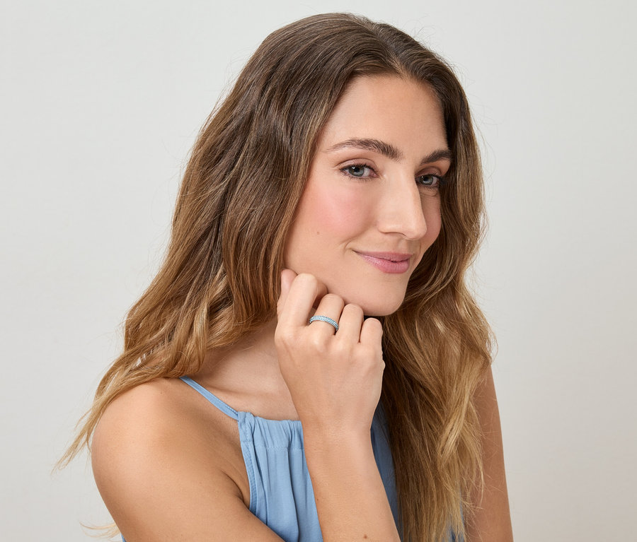 Kobieta z falowanymi włosami i w niebieskim topie dotyka palca z pierścionkiem na jasnym tle.