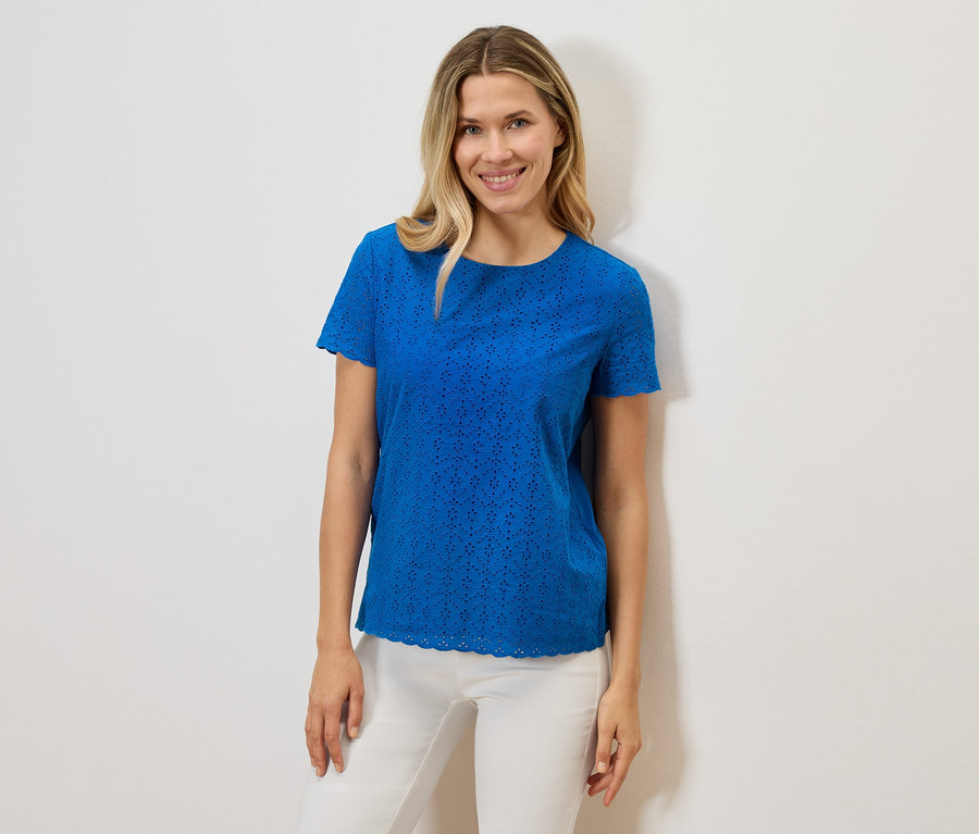 Uśmiechnięta kobieta ma na sobie niebieską, ażurową koszulkę z krótkim rękawem i białe spodnie na białym tle.