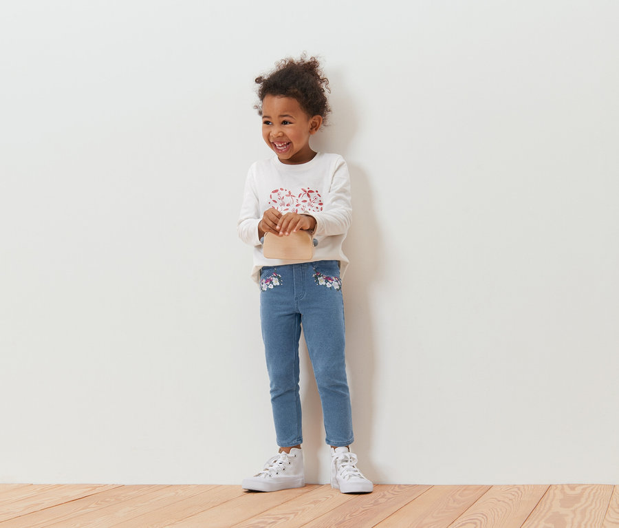 Uśmiechnięta, kręcona dziewczynka trzymająca beżową torebkę, w bluzce z długim rękawem i niebieskich dżinsach z kwiatowym haftem, stojąca na tle białej ściany.