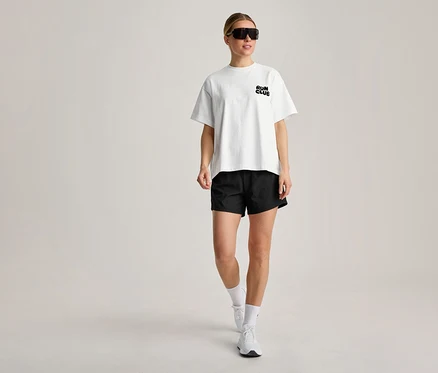 Kobieta idzie, ubrana w białą koszulkę sportową z napisem „RUN CLUB”, czarne szorty, białe skarpetki, białe sneakersy i czarne okulary przeciwsłoneczne.