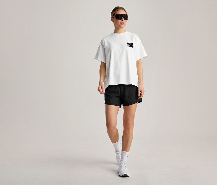Kobieta idzie, ubrana w białą koszulkę sportową z napisem „RUN CLUB”, czarne szorty, białe skarpetki, białe sneakersy i czarne okulary przeciwsłoneczne.