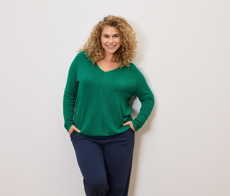 Kobieta z kręconymi włosami w zielonym swetrze i granatowych spodniach, z rękami w kieszeniach.
