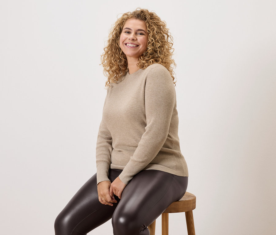 Kobieta z kręconymi włosami siedzi na drewnianym stołku, ubrana w beżowy sweter i ciemne legginsy.