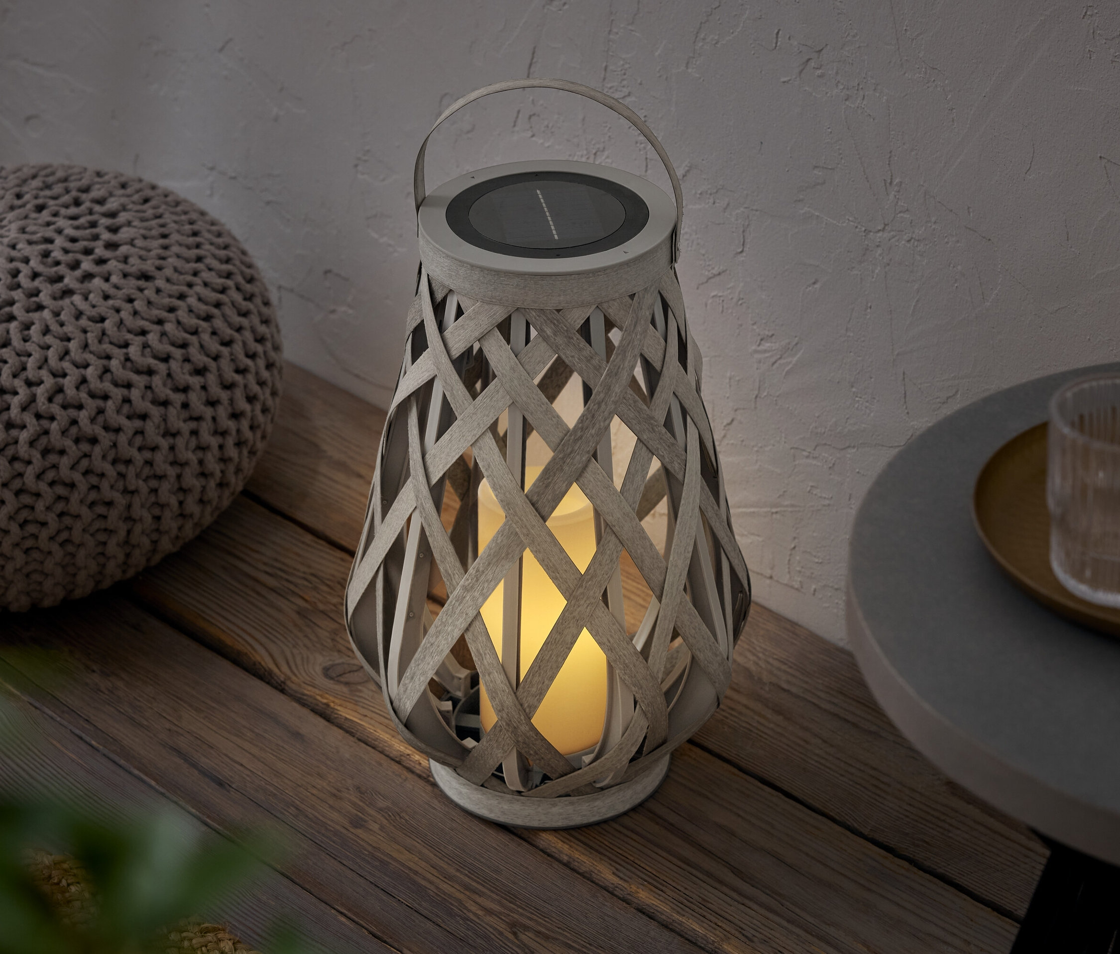 Pleciony lampion z panelem słonecznym stoi obok pufa na drewnianej podłodze.