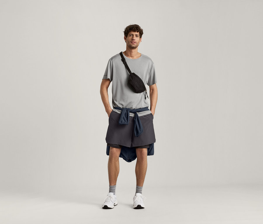 Mężczyzna ma na sobie jasno szarą koszulkę sportową, czarną torbę na biodra, szorty i kurtkę zawiązaną wokół bioder.