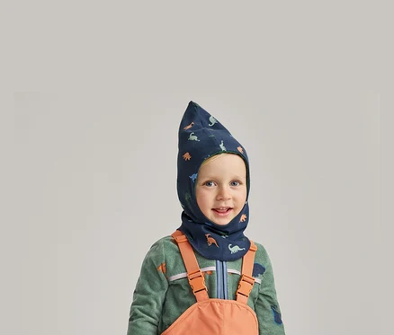 Uśmiechnięty chłopiec w niebieskiej kominiarce w dinozaury, zielonej bluzie polarowej i pomarańczowych ogrodniczkach na szarym tle.