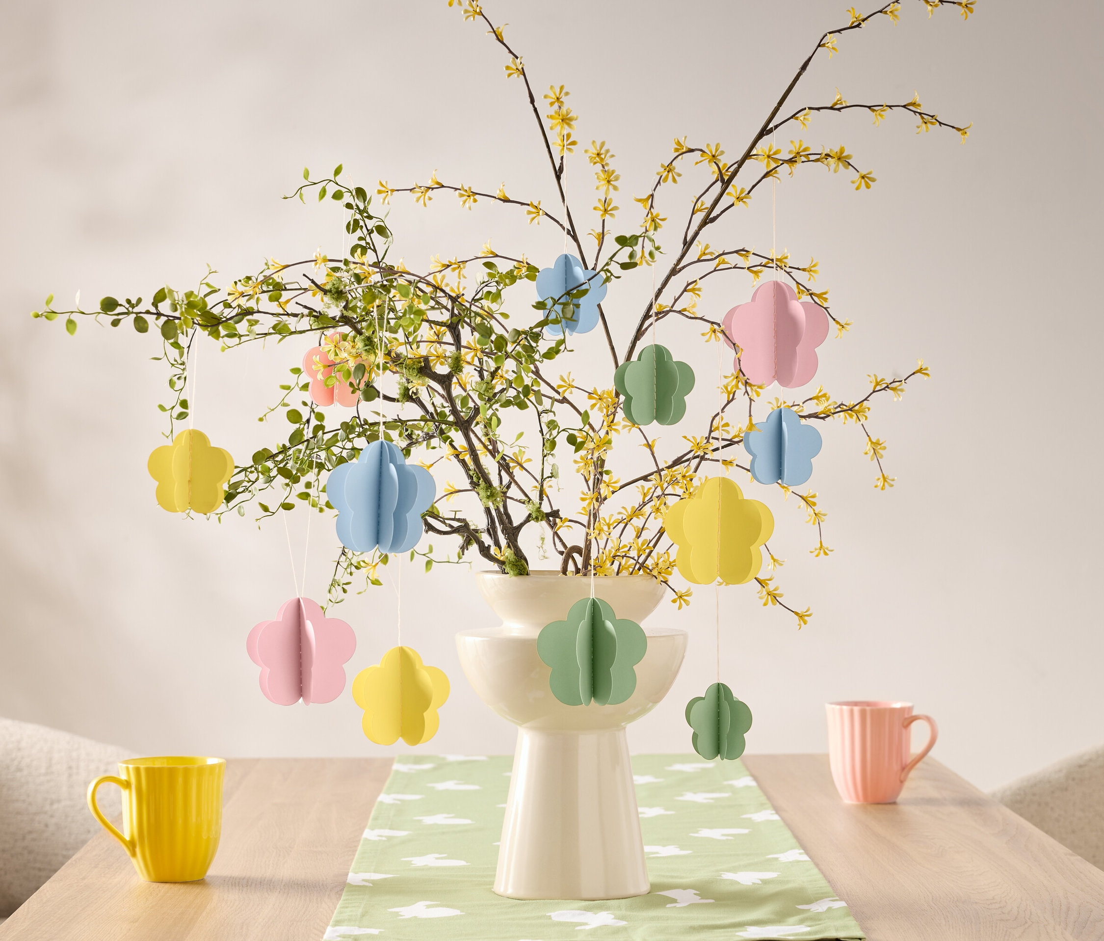 Kwiatowe ozdoby wiszą na gałęzi w wazonie na stole, obok żółtego i różowego kubka.