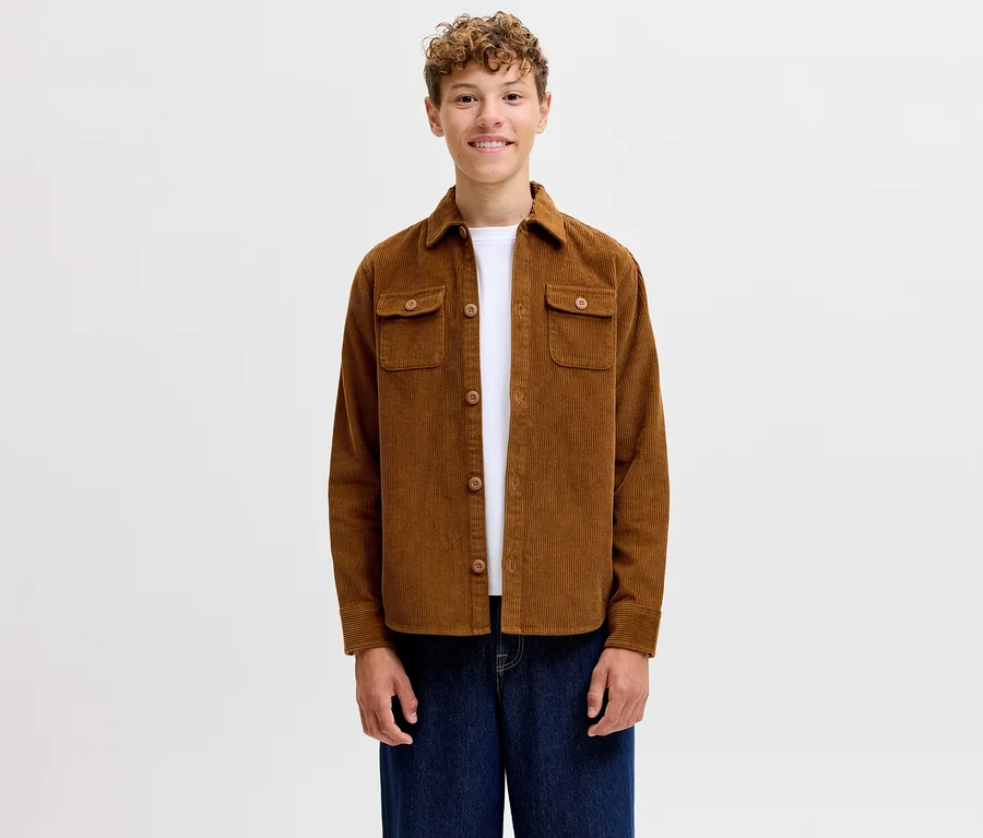 Uśmiechnięty młody mężczyzna w brązowej koszuli sztruksowej Jack&Jones Regent, białym T-shircie i niebieskich dżinsach.