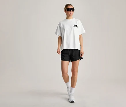 Kobieta idzie, ubrana w białą koszulkę sportową z napisem „RUN CLUB”, czarne szorty, białe skarpetki, białe sneakersy i czarne okulary przeciwsłoneczne.