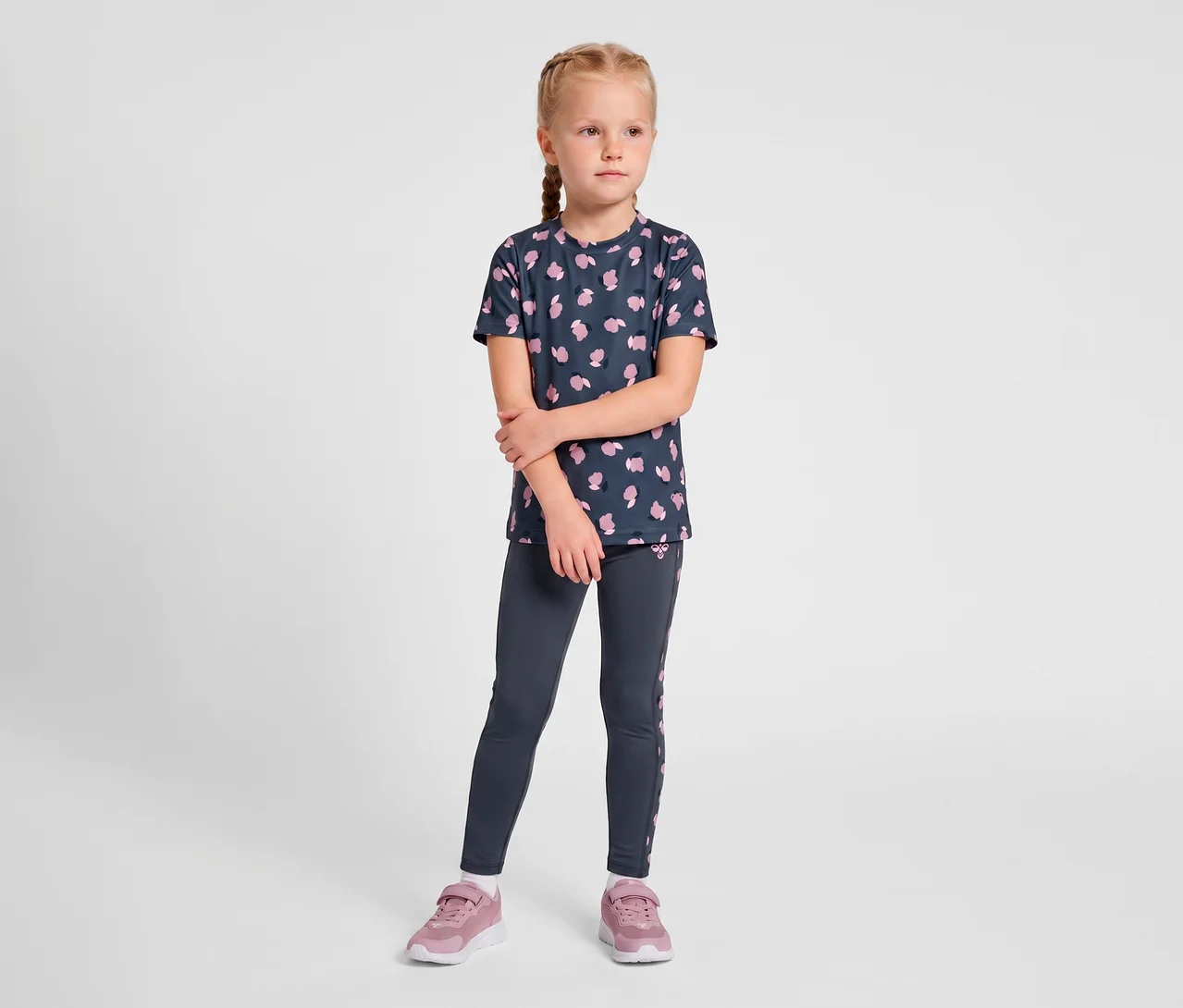 Młoda dziewczynka z zaplecionymi blond włosami stoi, ubrana w ciemnoniebieską koszulkę z różowymi wzorami, ciemnoniebieskie legginsy hummel® „HMLJR AOP Gym Tights” z różowymi paskami po bokach i różowe tenisówki.