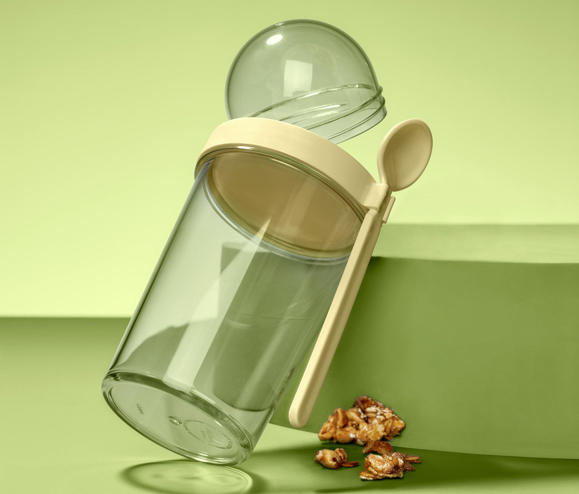 Przezroczysty kubek na jogurt i musli z beżową pokrywką, łyżką i przezroczystym pojemnikiem kulkowym stoi pochylony na zielonej powierzchni obok kawałków musli.