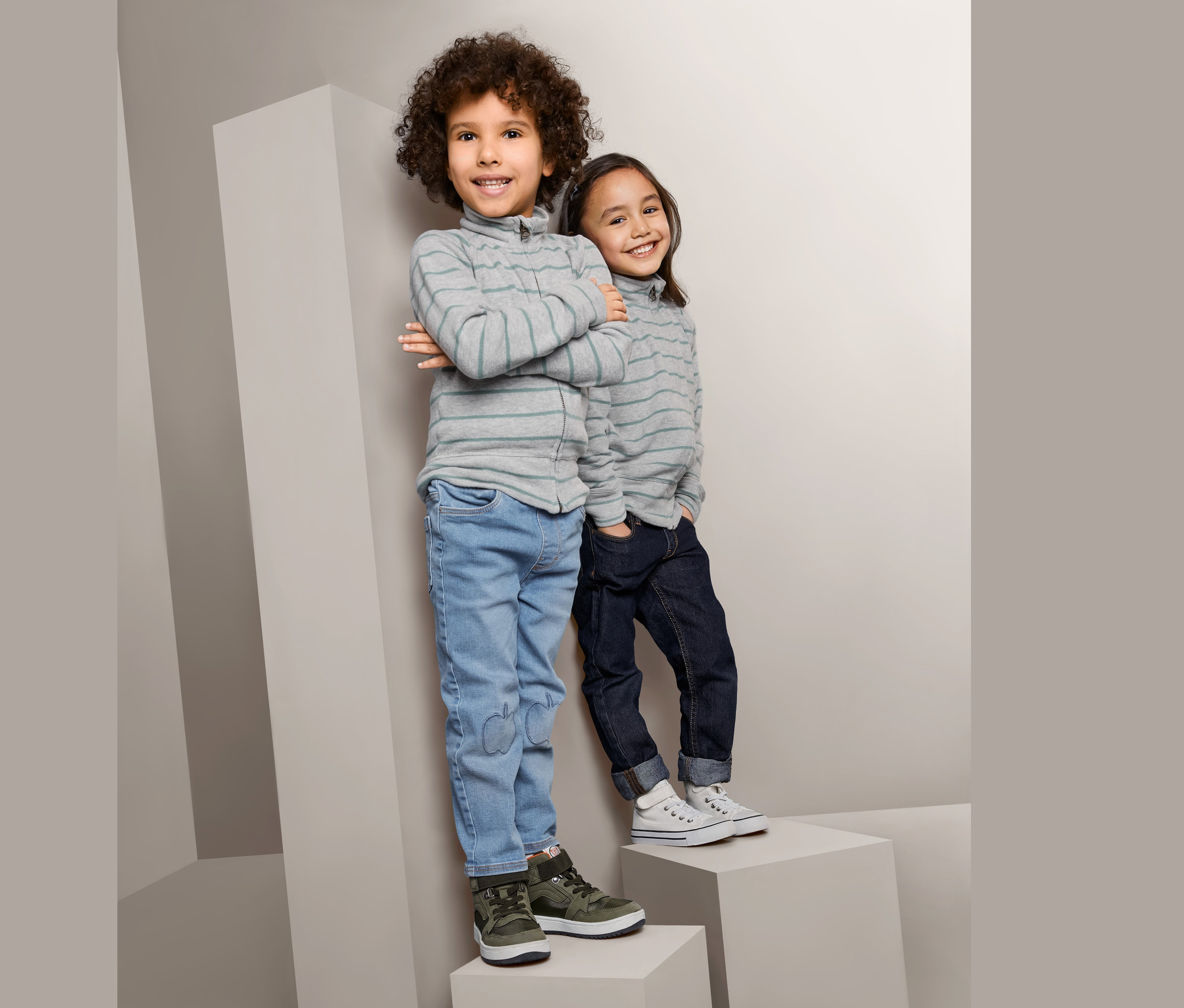 Dwoje dzieci stoi na szarych blokach. Chłopiec po lewej stronie ma na sobie Dziecięcy, rozpinany sweter z dzianiny w sportowym stylu, dżinsy i zielone buty. Dziewczynka po prawej stronie ma na sobie Dziecięcy, rozpinany sweter z dzianiny w sportowym stylu, ciemne dżinsy i białe buty.