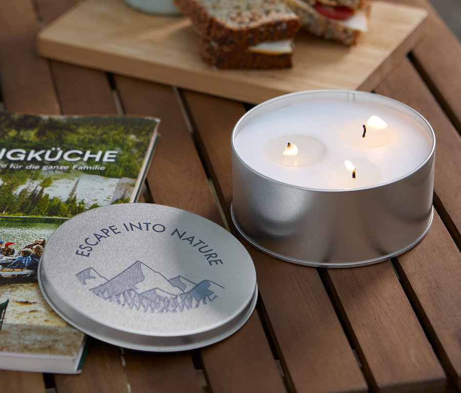 Na drewnianym stole leżą świeca citronella, książka Campingküche, kubek i kanapki.