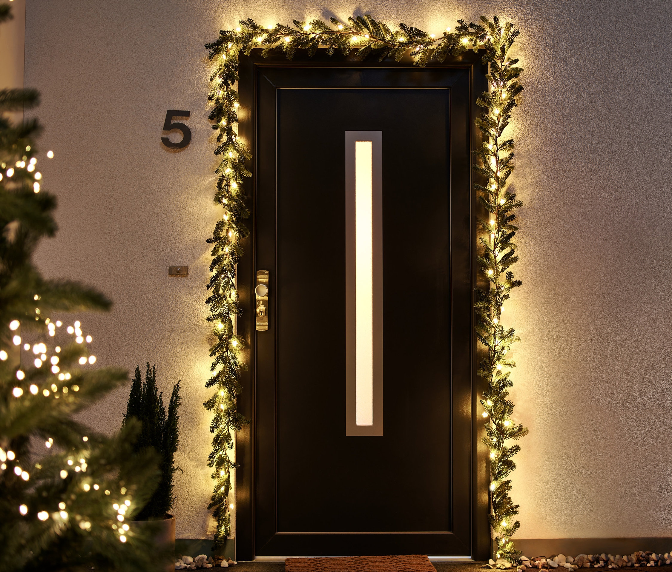 Drzwi wejściowe ozdobione girlandą choinkową LED, po lewej stronie choinka z lampkami LED.