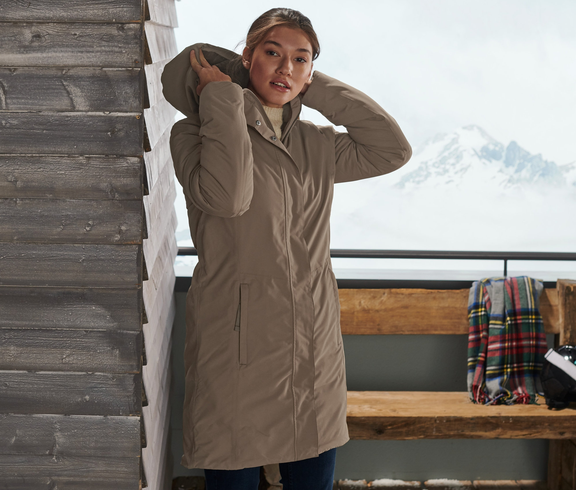 Kobieta poprawia kaptur szarobeżowego płaszcza ciepłochronnego. W tle widoczne narty i ośnieżony krajobraz.