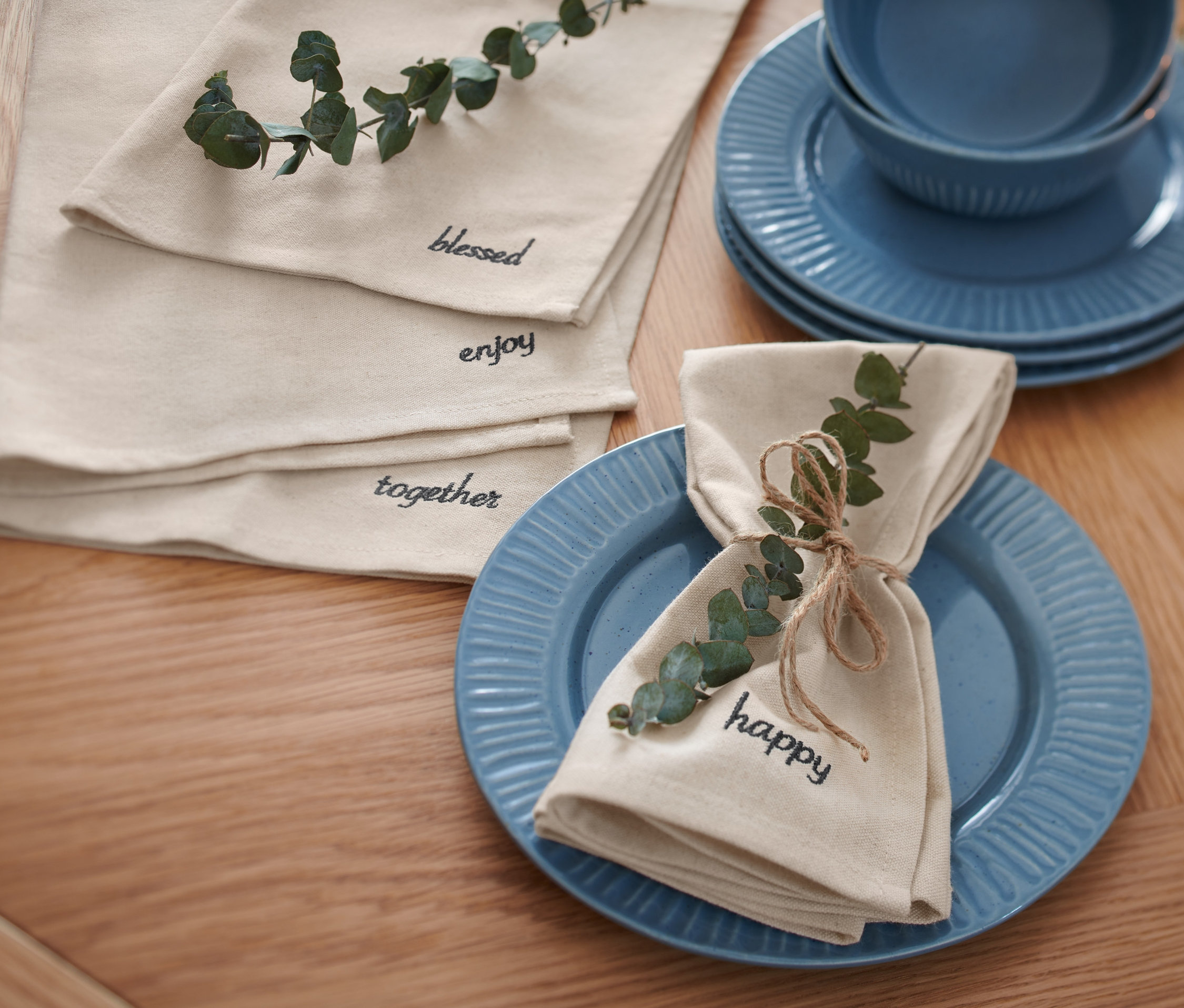 Na drewnianym stole leżą niebieskie talerze i złożone beżowe serwetki z napisami i eukaliptusem. Na górze leżą Haftowane serwetki, 4 sztuki.