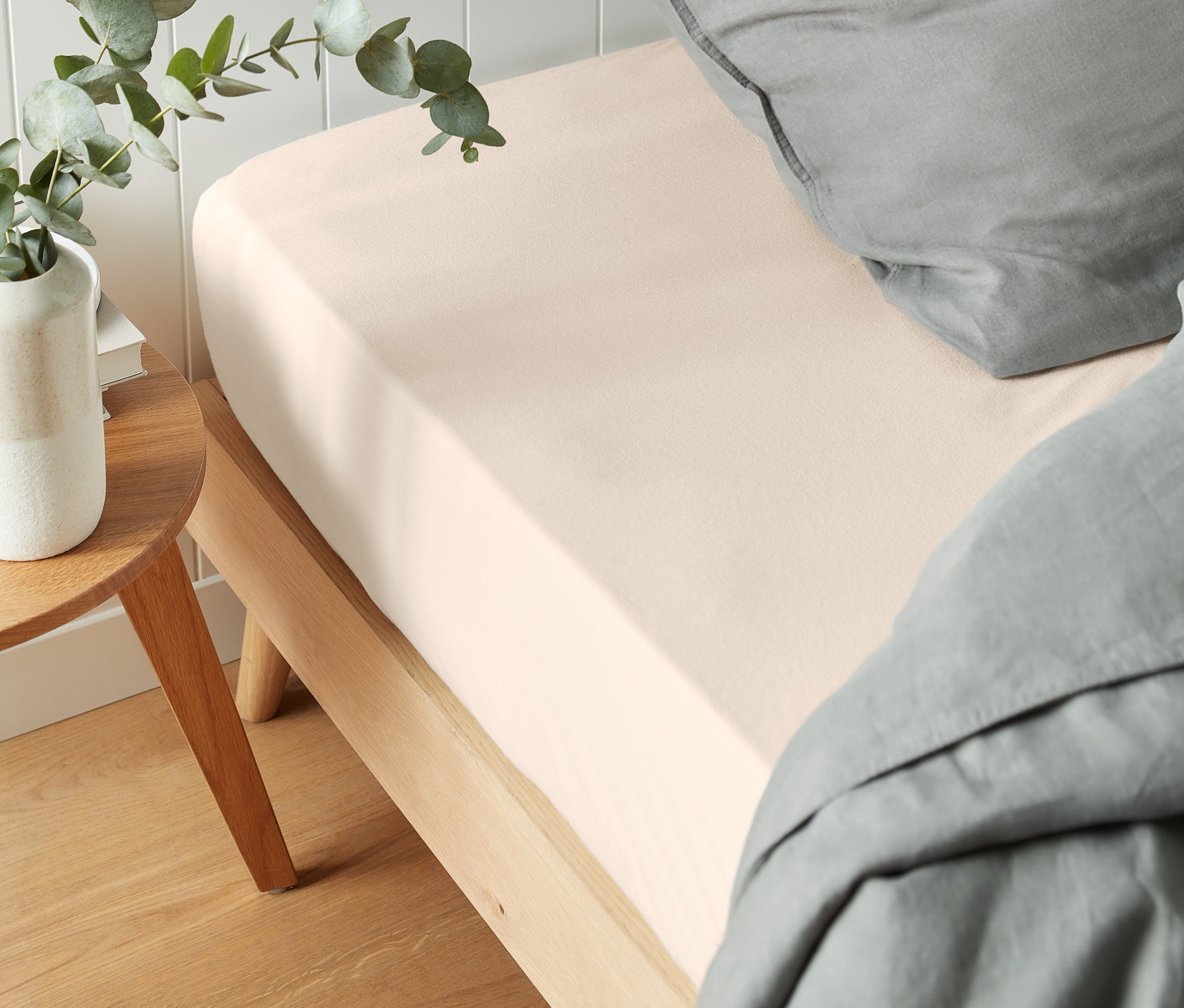 Jasne prześcieradło flanelowe z gumką na łóżku z szarą pościelą i rośliną na szafce nocnej.