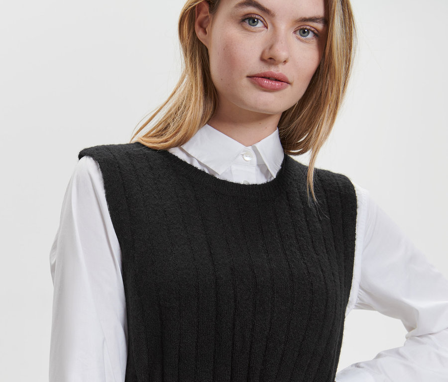 Blondynka ma na sobie czarną sukienkę z prążkowanej dzianiny i białą koszulę.