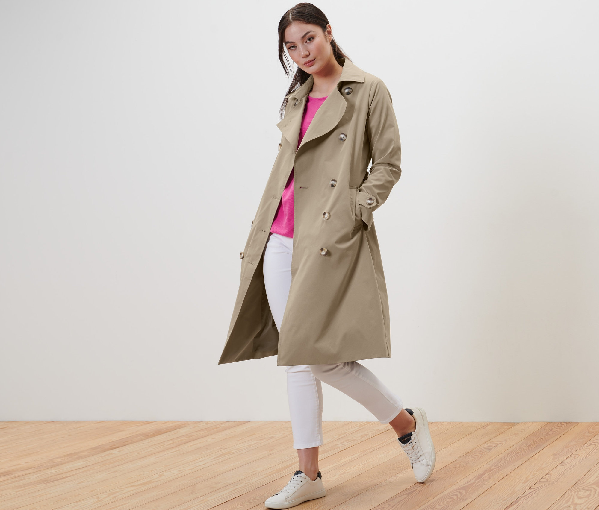 Kobieta pozuje w beżowym prochowcu, białych dżinsach i butach sportowych.