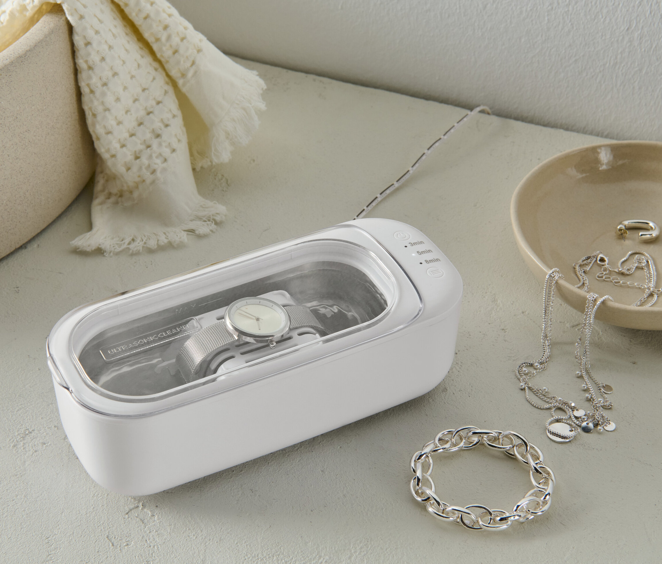 Zegarek czyszczony w białym ultradźwiękowym urządzeniu oczyszczającym. Obok leży biżuteria.