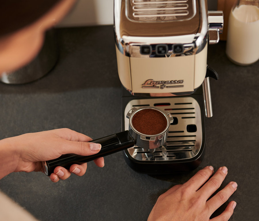 Kobieta trzyma kolbę z kawą nad ociekaczem kremowego ekspresu kolbowego do kawy »Lapressa«.