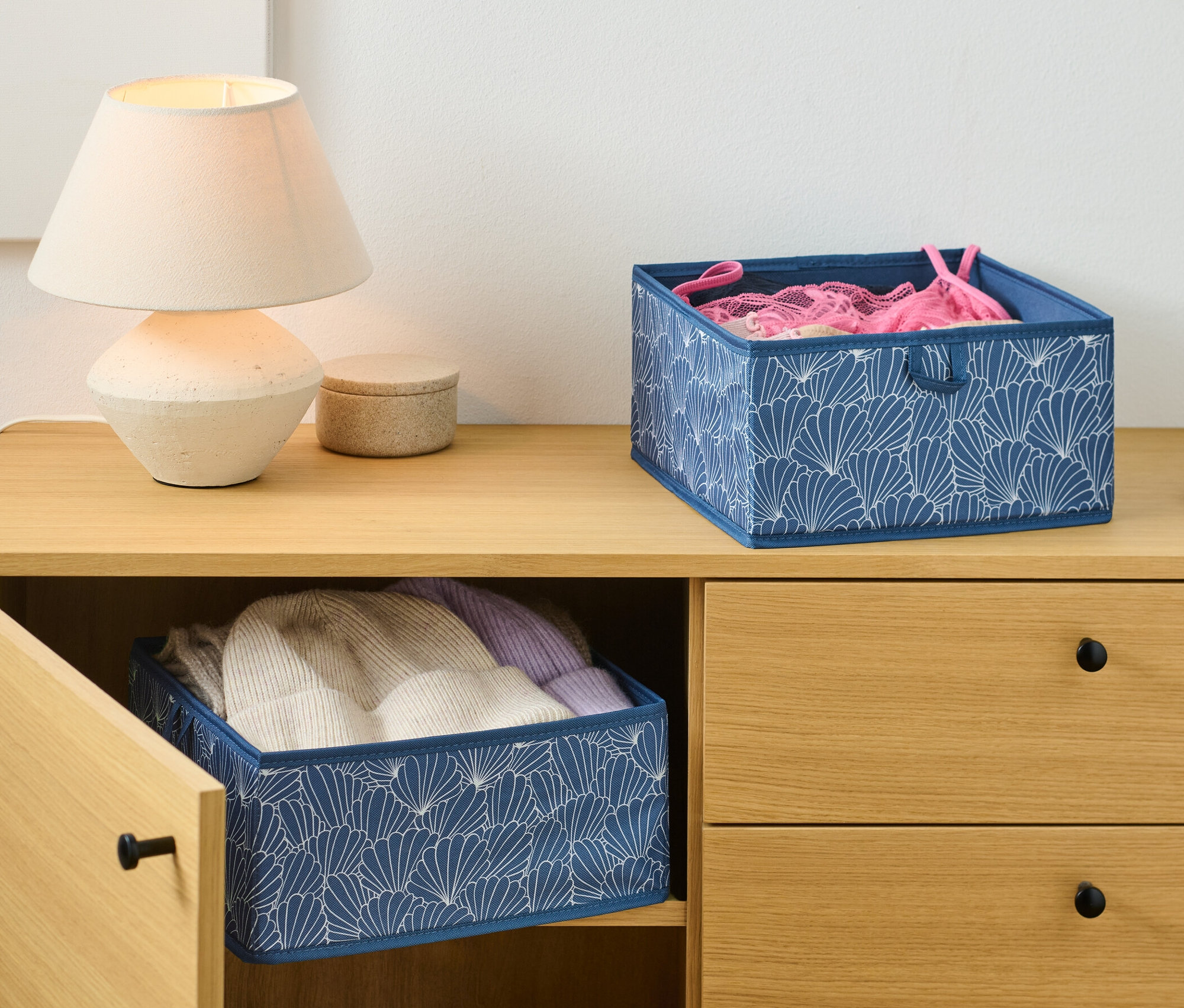 Na komodzie stoi lampa i dwa duże organizery do szuflad, jeden z ubraniami w środku.