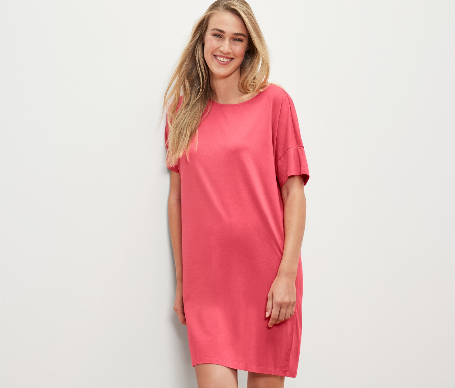 Blondynka w koralowej koszuli nocnej opiera się o białą ścianę.