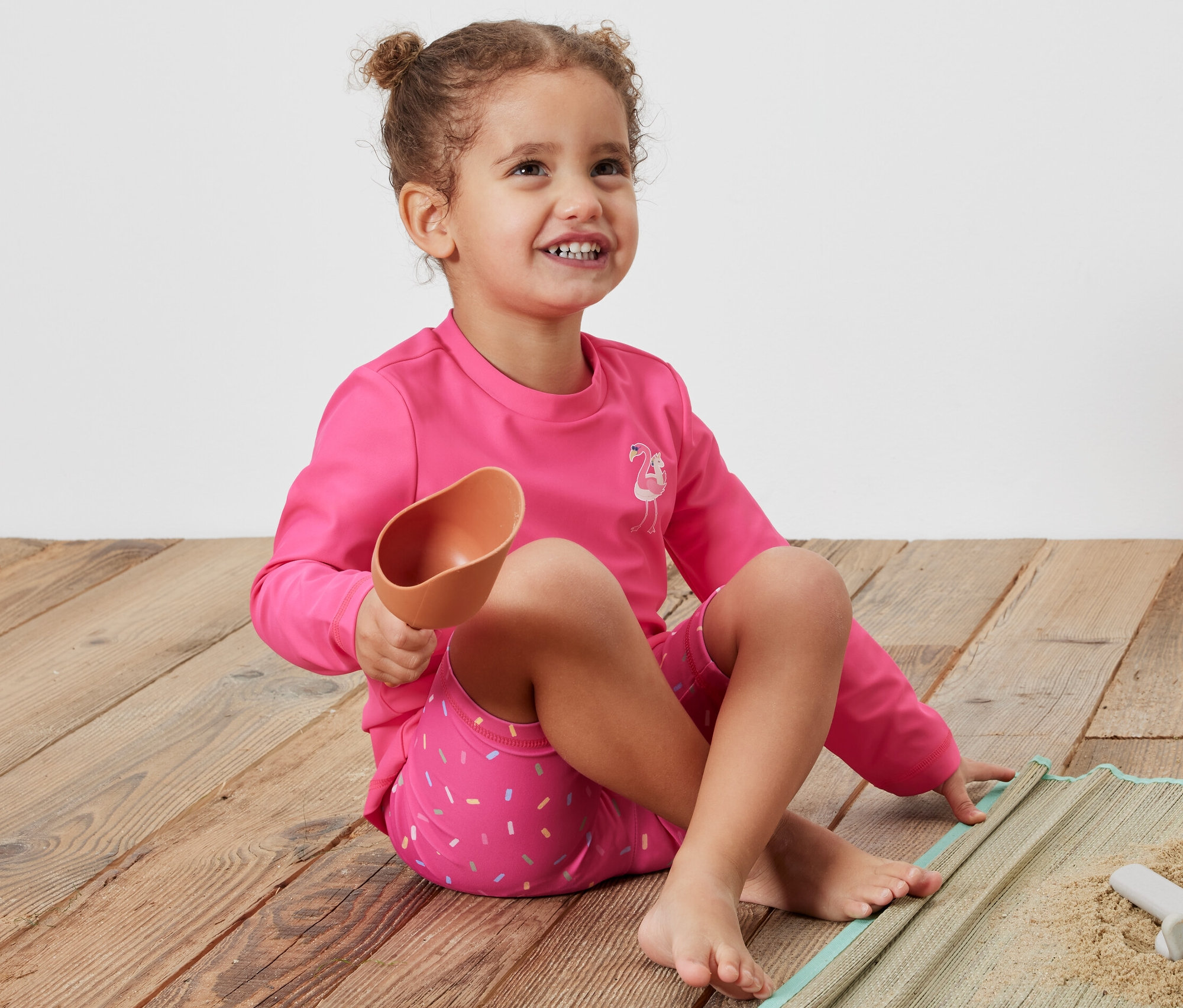 Dziewczynka siedzi na kocu i trzyma łopatkę, ubrana w różowy komplet z filtrem UV 80.