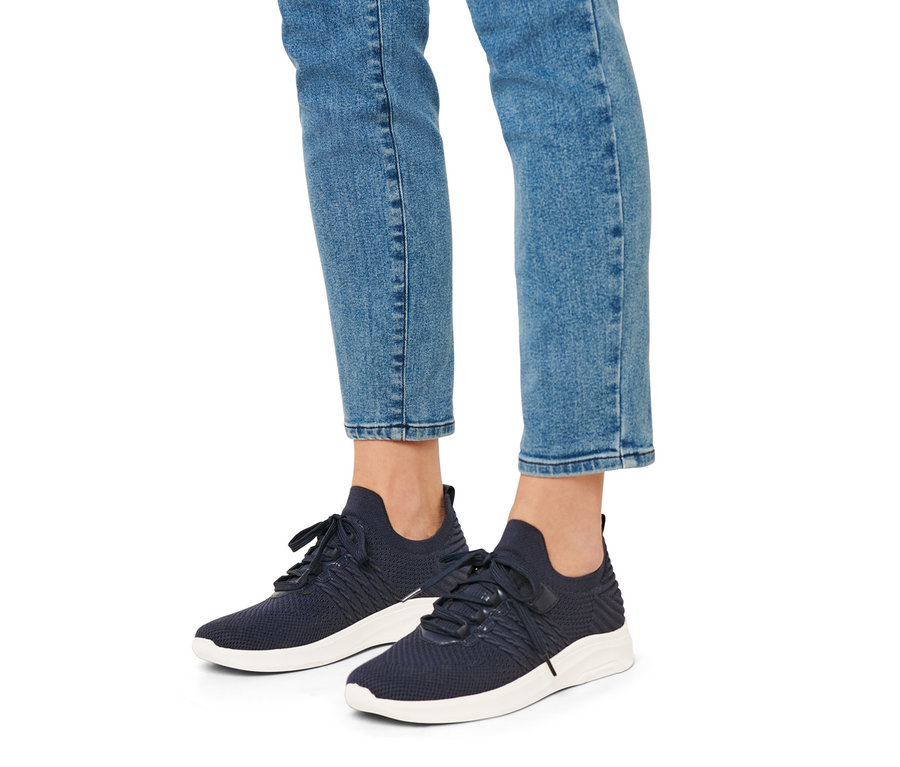 Nogi osoby w jeansach i niebieskich wsuwanych butach typu sneaker z białymi podeszwami.