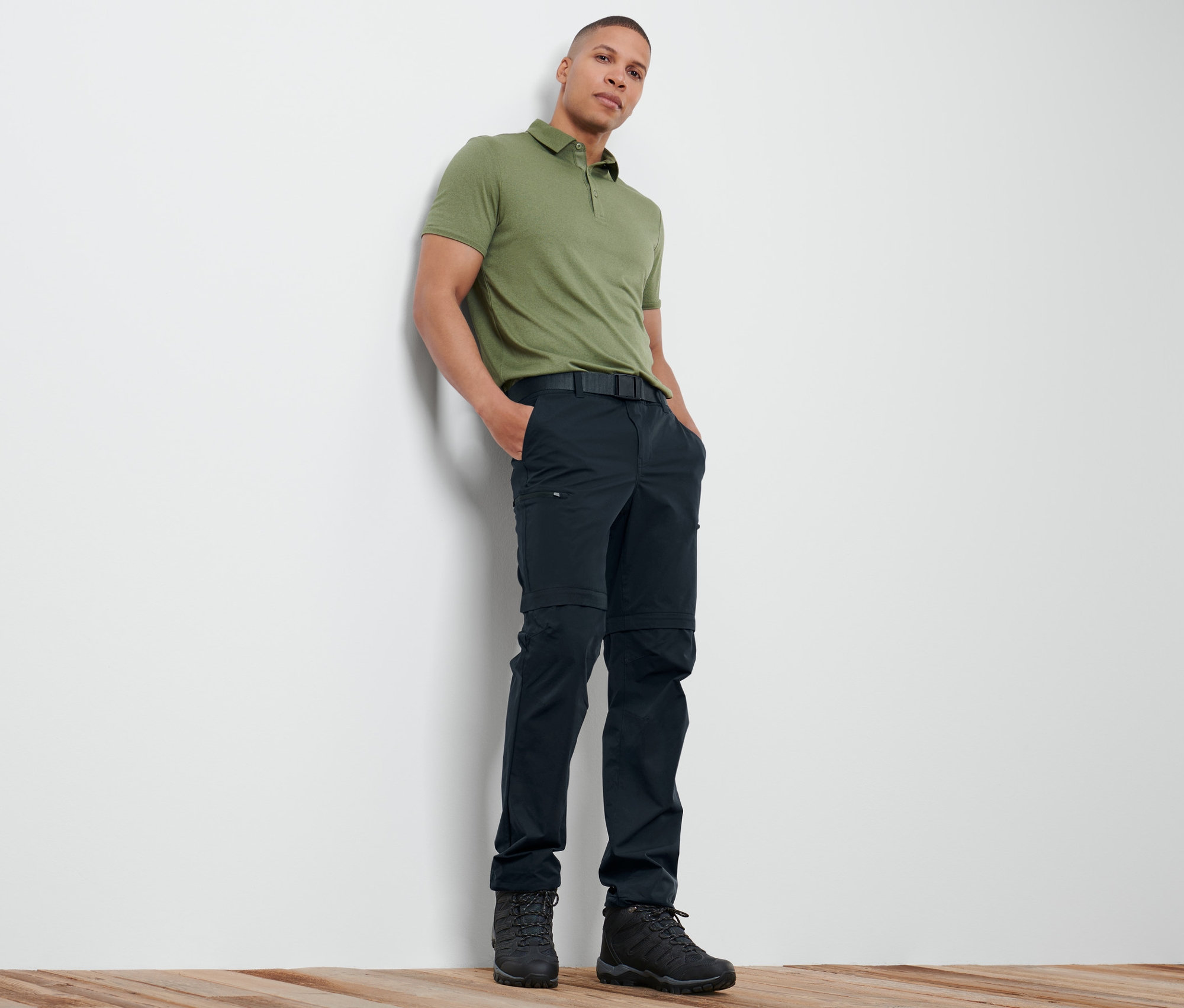 Mężczyzna oparty o ścianę ma na sobie zieloną koszulkę polo i granatowe spodnie funkcyjne 2 w 1.