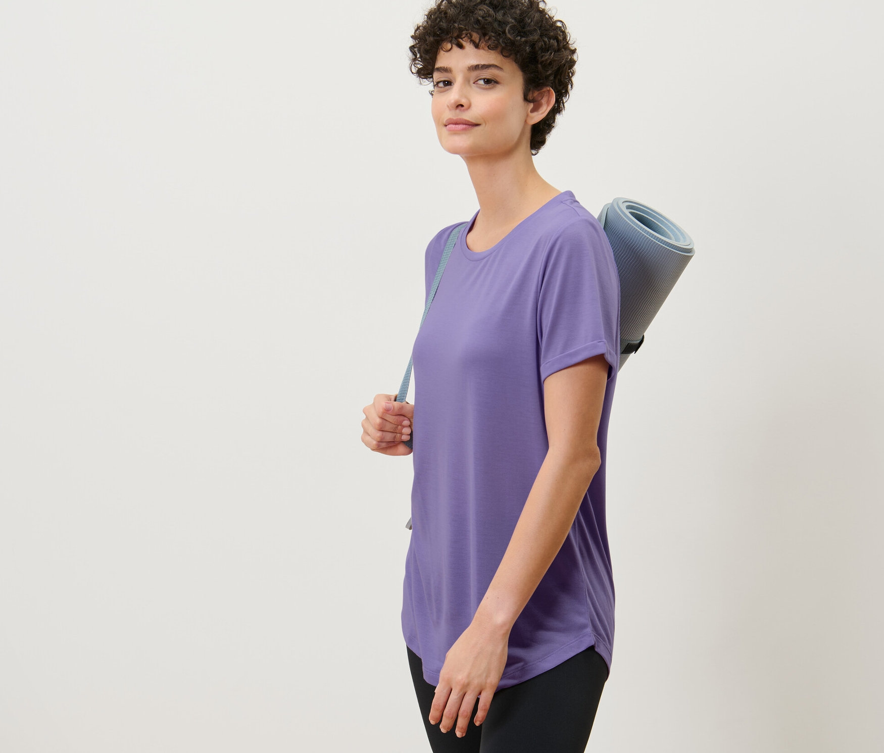 Kobieta ma na sobie liliową długą koszulkę i niesie matę do ćwiczeń.