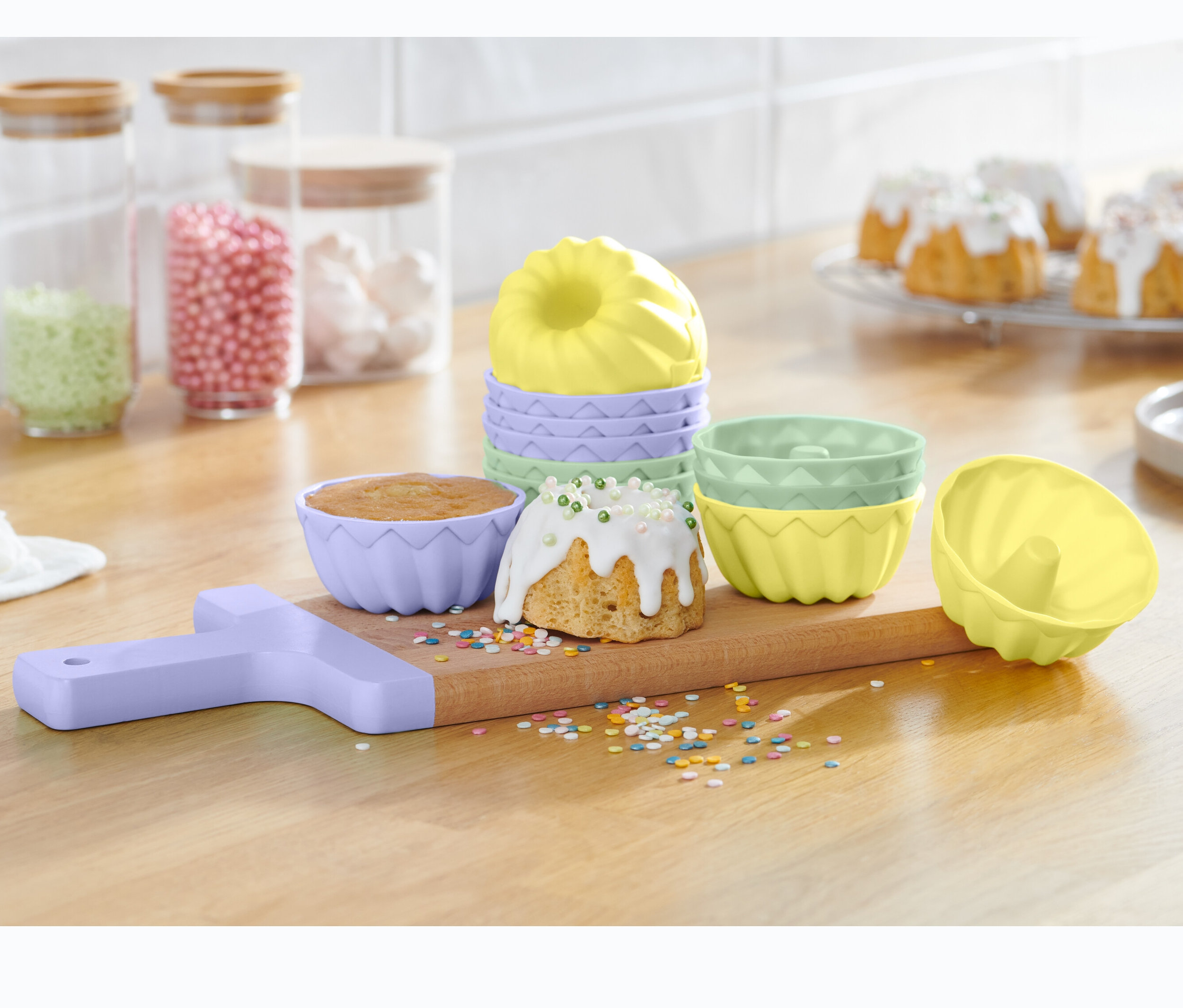 Na drewnianej desce znajdują się foremki do minibabeczek, 12 sztuk, wraz z upieczonymi babeczkami i posypką.