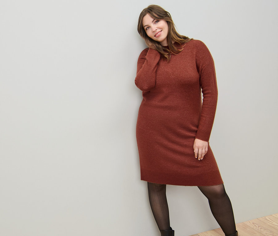 Kobieta opiera się o ścianę, ubrana w dzianinową sukienkę z zawartością wełny i cienkie rajstopy 50 DEN z efektem przezroczystości.