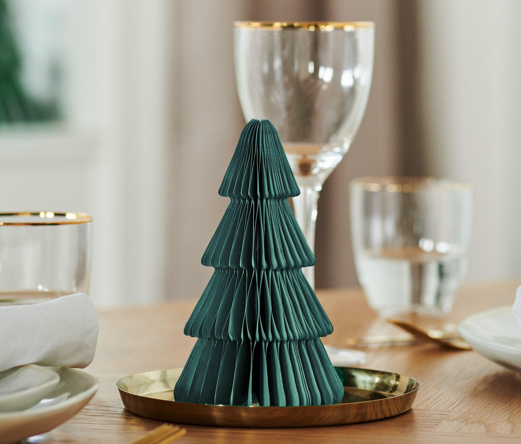 Mała, ciemnozielona składana choinka z papieru na złotej tacy stoi na nakrytym stole.