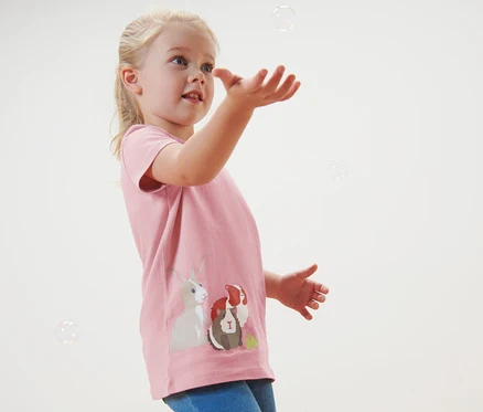 Dziewczynka łapie bańki mydlane. Ma na sobie różowy t-shirt z obrazkami zwierząt. Koszulki, 2 sztuki.