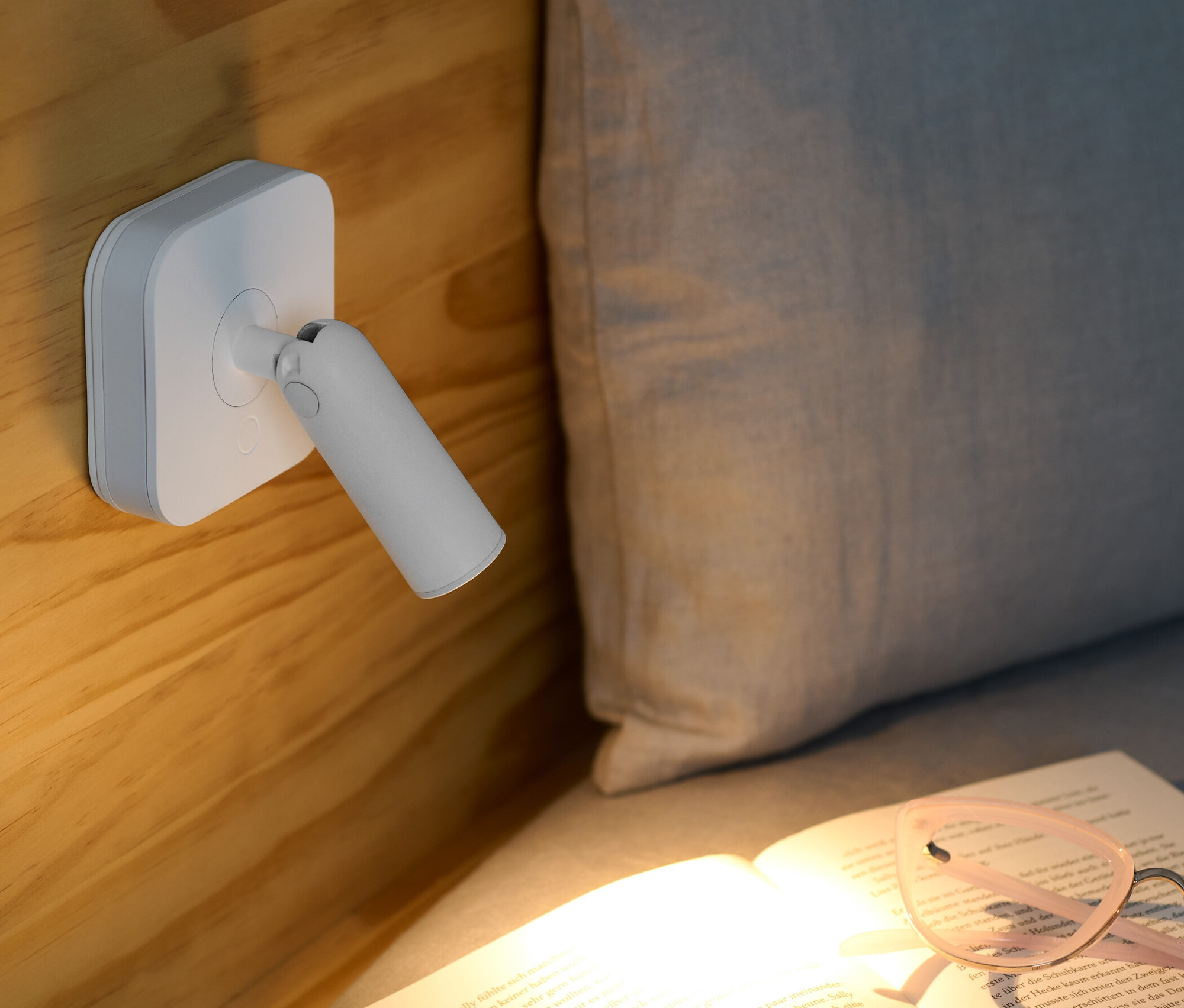 Lampka nocna LED z reflektorem przymocowana jest do drewnianego łóżka i oświetla otwartą książkę z okularami.