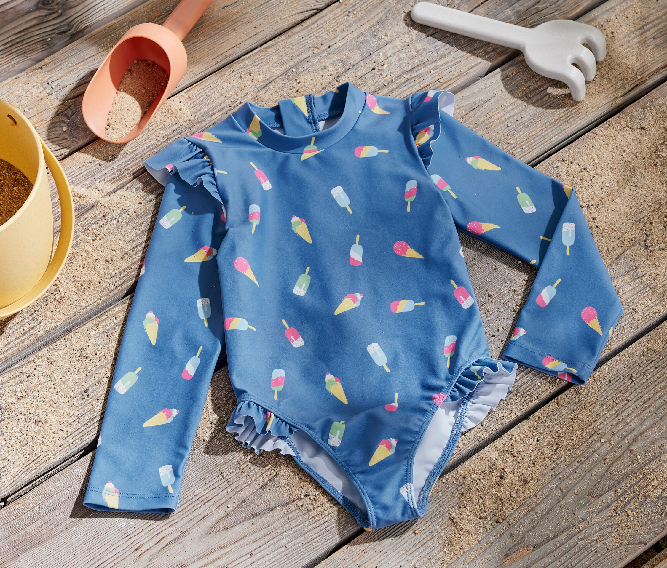 Niebieski kostium kąpielowy dla dzieci z nadrukiem lodów i filtrem UV 80 leży na drewnianej desce obok zabawki do piasku.