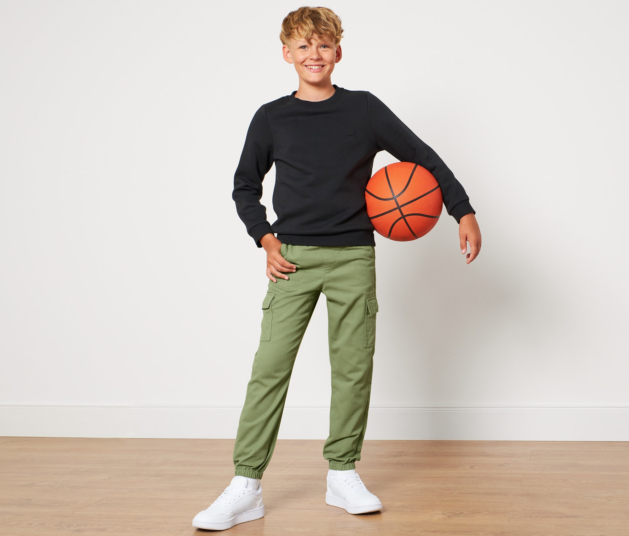 Chłopiec pozuje z piłką do koszykówki, ubrany w spodnie bojówki na gumce.