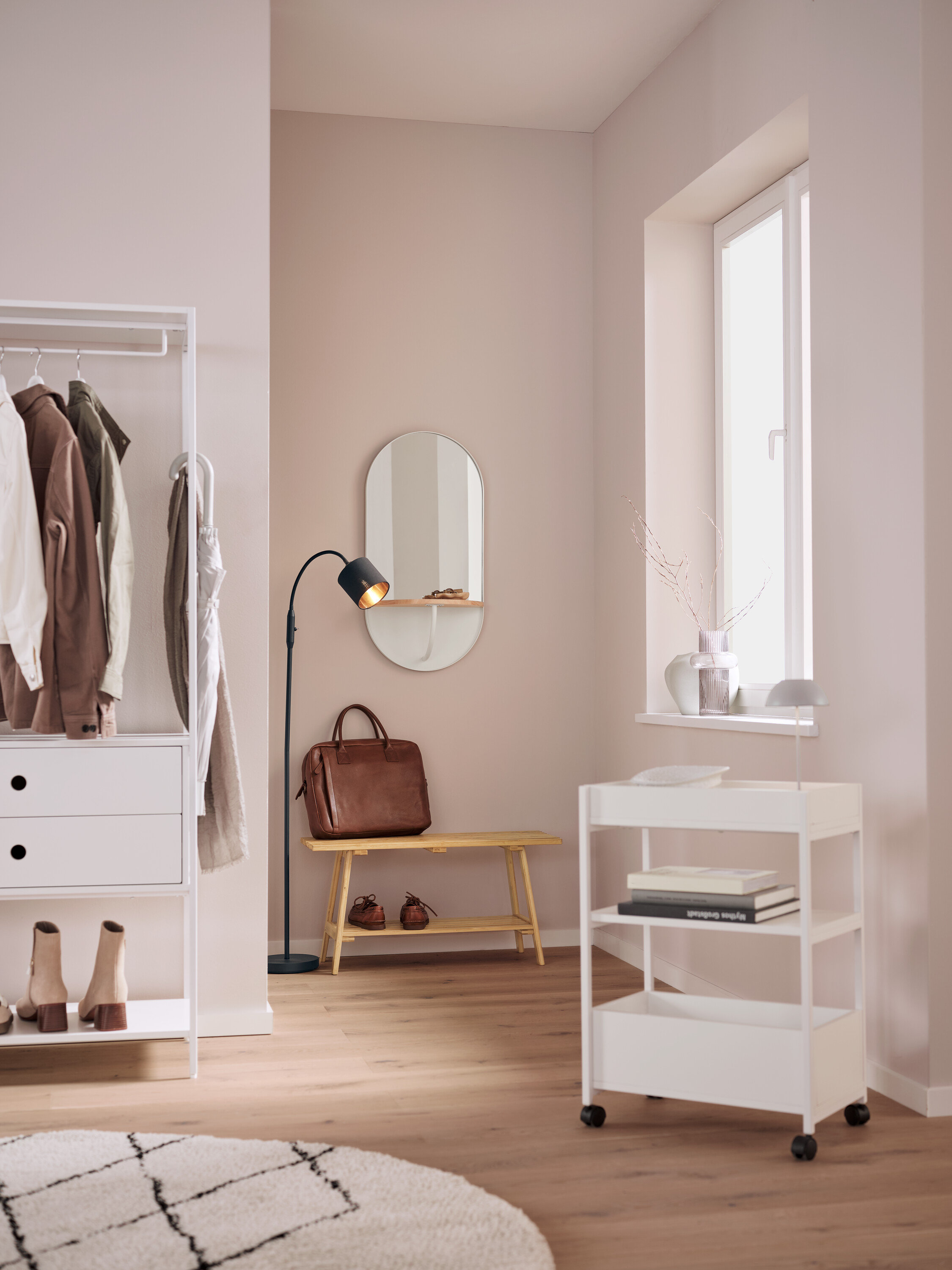 We wnętrzu znajduje się biały regał z ubraniami, owalne lustro, brązowa skórzana torba na ławce, czarna lampa i biały wózek na kółkach.
