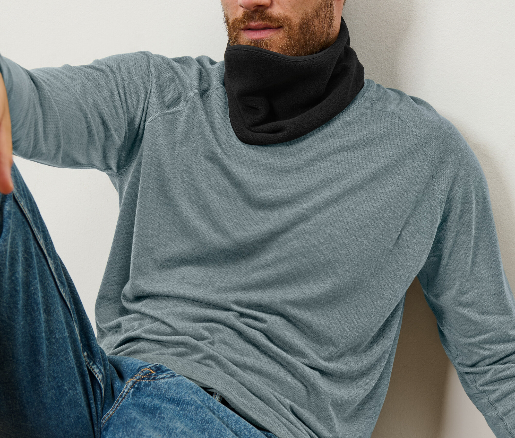 Mężczyzna siedzi oparty o ścianę, ma na sobie czarny ocieplacz na szyję, niebieskie dżinsy i niebiesko-szarą koszulkę z długim rękawem.