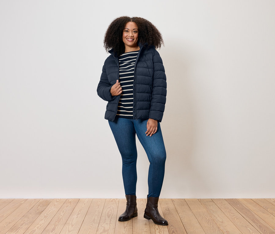 Kobieta z kręconymi włosami stoi i ma na sobie niebieską kurtkę pikowaną, koszulkę w paski, dżinsy i buty.