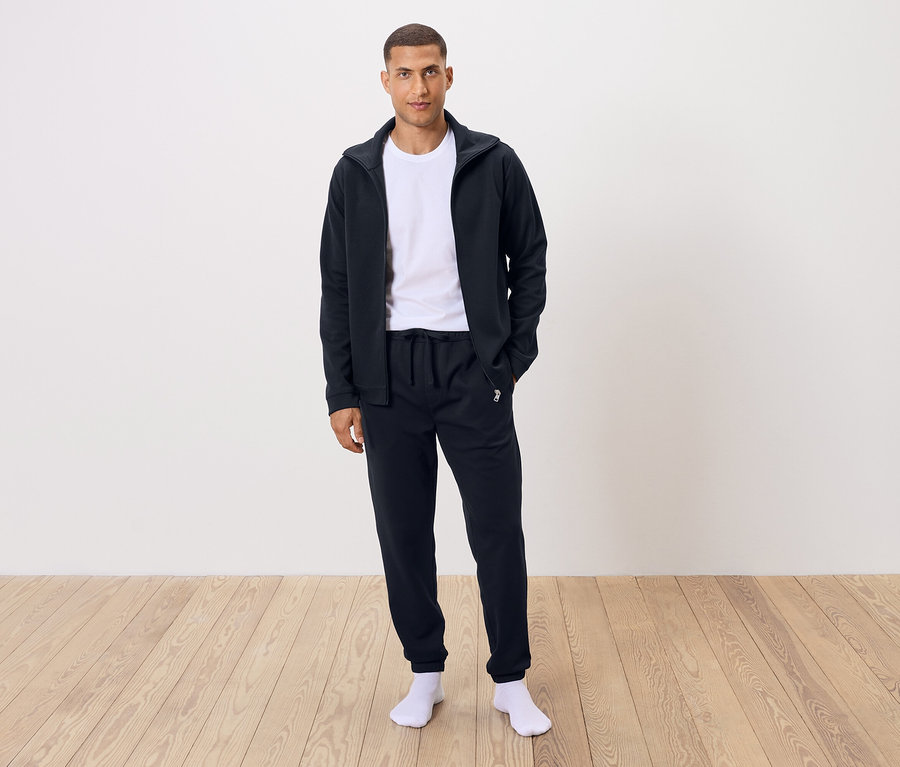 Mężczyzna stoi ubrany w białą koszulkę, czarne spodnie dresowe i czarną bluzę na zamek.